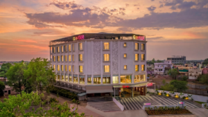 Paved driveway in foreground leading to a multi-storey white hotel building with neon signage under a sunset sky.