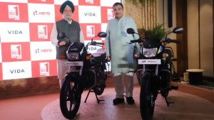 Indian ministers standing with two black Hero motorcycles, Splendor+ and HF Deluxe, on an indoor stage with Hero and VIDA logos.