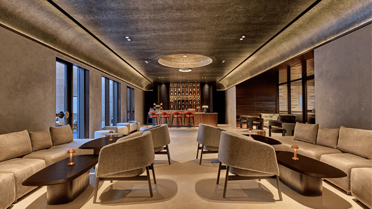 The 15-acre beachfront resort for your next break 3 A sophisticated indoor hotel lounge with plush grey seating, low wooden tables, a central circular ceiling light, and a fully stocked bar.