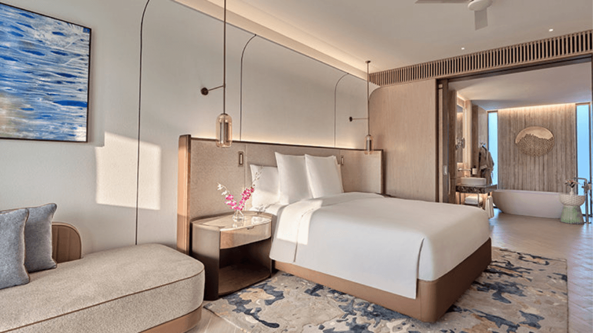 The 15-acre beachfront resort for your next break 2 A modern resort bedroom with a white king bed, beige accent walls, blue carpet pattern, and an open bathroom with a standalone bathtub.