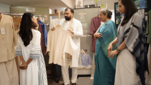 A man presents a cream top to three women by a Khadi India Swadha banner at a wellness apparel showcase.