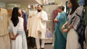 A man presents a cream top to three women by a Khadi India Swadha banner at a wellness apparel showcase.