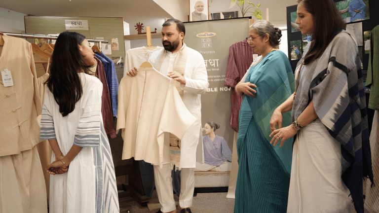 With 9 gender-neutral styles, this collection rethinks yoga 7 A man presents a cream top to three women by a Khadi India Swadha banner at a wellness apparel showcase.