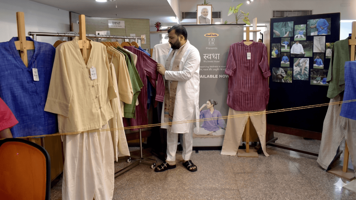 A man inspects a striped maroon shirt on a clothing rack next to a Khadi India Swadha exhibition display.
