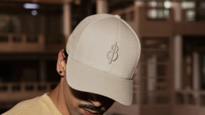 A man wearing a light beige cap with an embroidered logo, looking down against a blurred background, showcasing sportswear.