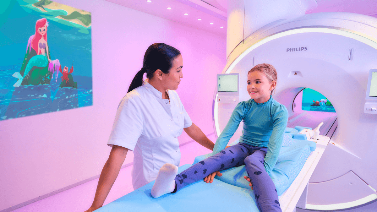 A medical worker talks to a smiling girl on a Philips MRI bed under pink lights with a Little Mermaid wall projection.