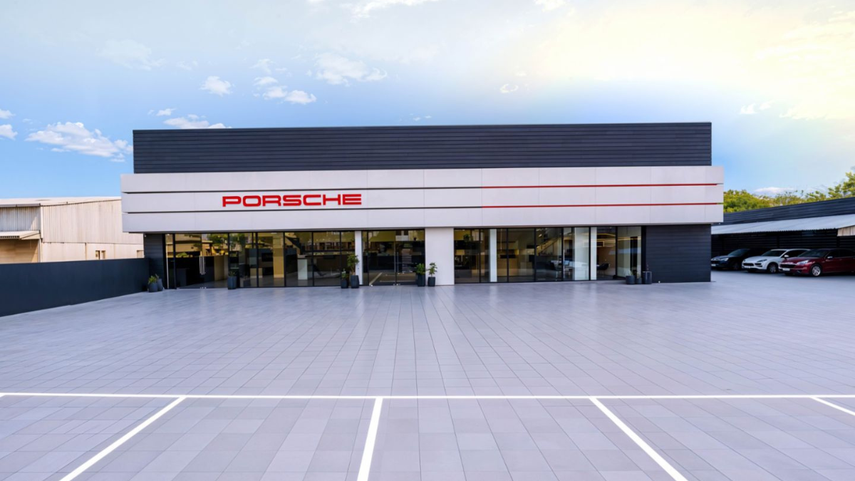 A wide exterior shot of a modern grey and white building with a red Porsche logo facing a tiled forecourt.
