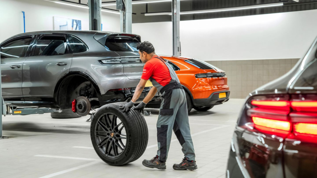 A technician rolling a car tyre next to a grey SUV on a mechanical lift inside a brightly lit workshop.