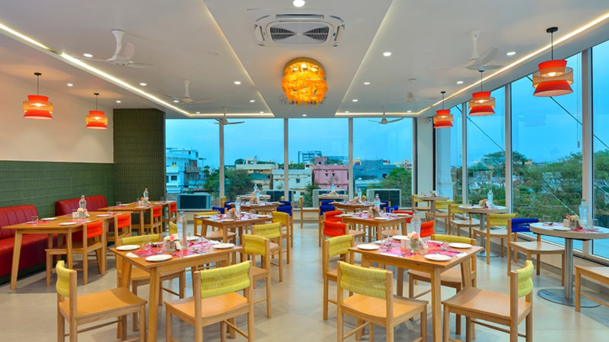At ₹3,399 a night, IHCL brings lean luxury to Gadchiroli 2 Wooden dining tables and yellow chairs inside a brightly lit restaurant with large glass windows showing a town.