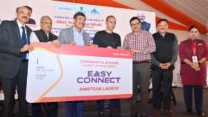 Officials hold an Air India Easy Connect Amritsar board with a passenger on stage in front of event backdrop.