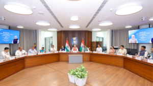 Seated officials around a wooden table flanked by digital screens and Indian flags at a Delhi briefing.