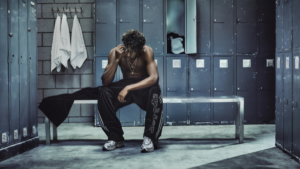 A man sits on a bench wearing black trackpants and silver sneakers inside a blue locker room with towels on the wall.