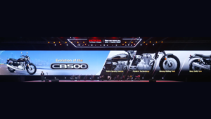 Presenter on stage beside a wide digital display presenting the Honda CB500 roadster motorcycle graphics.