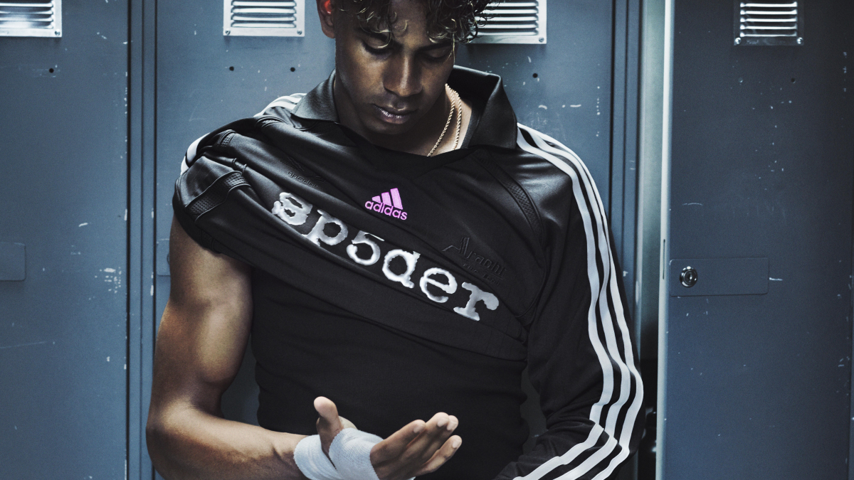 Can a punk streetwear label redesign an iconic football shoe? 2 A man with a wrapped wrist looks down while adjusting a black jersey with pink branding in front of blue metal lockers.