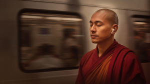 A man in red and orange robes stands with closed eyes wearing a cream earbud, with a blurred train moving in the background.