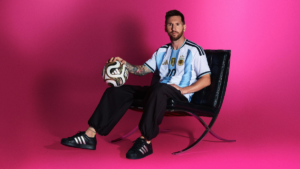Lionel Messi sits in a black armchair holding a gold and black football against a bright pink studio backdrop.