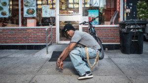 A man crouching on a pavement in a beanie and loose jeans, wearing black and white Puma Suede 94 trainers.