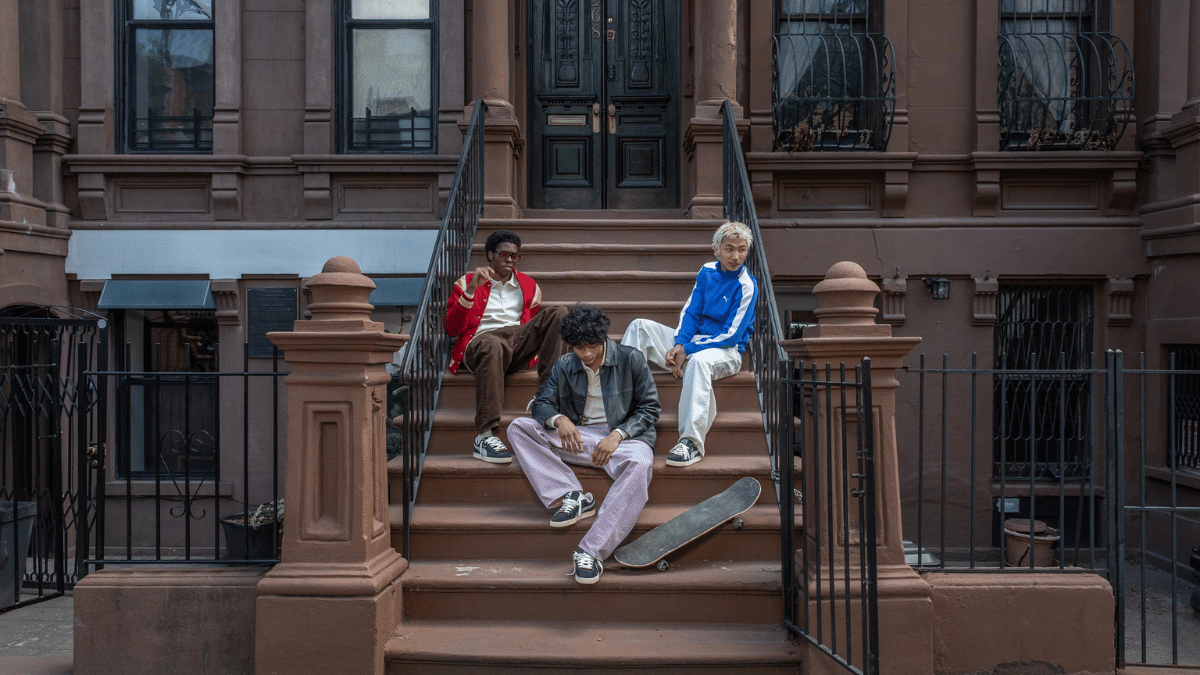 Three people wearing streetwear sit on brownstone building steps next to a skateboard, wearing lifestyle trainers.