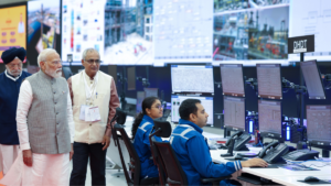 Narendra Modi walks past operators in blue uniforms sitting before monitors in a refinery control room in India.