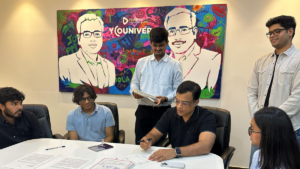 A man in a black shirt signs papers at a white conference table with colleagues in front of a corporate backdrop art piece.