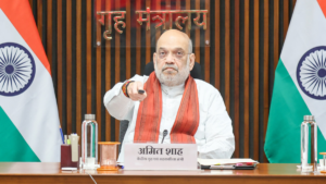 Amit Shah seated between two Indian flags pointing a remote control forward during the Sahakar Bhavan ceremony.