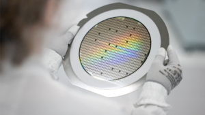 Gloved hands holding a circular semiconductor wafer with iridescent grid patterns in a lab for EV components.