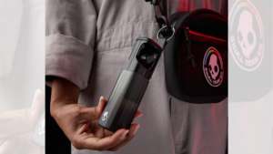 A hand holding a black wireless earbud slider case clipped onto a black shoulder bag strap with an O-ring.