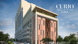 Multi-storey hotel building with balconies on left, pink exterior panels on right, and text overlay under a blue sky.