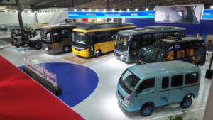 Blue mini-van and corporate passenger buses displayed on a brightly lit exhibition floor with branding signs in India.