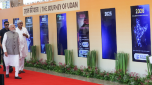Prime Minister Narendra Modi walks left to right on a red carpet past a wall with vertical UDAN scheme timeline panels.