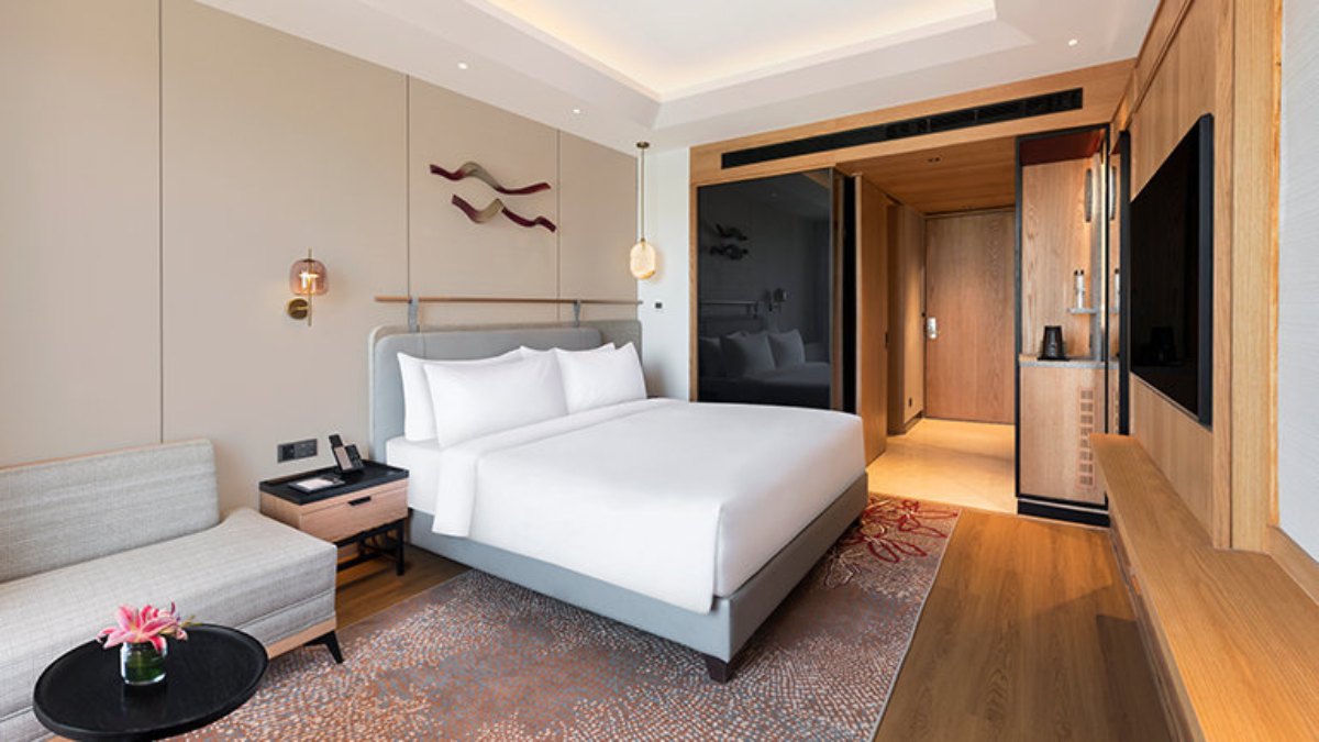 Panchkula gets India's first Vignette Collection hotel 2 Grey sofa on left foreground, white bed on patterned rug center, wooden wall with television right, guest room.
