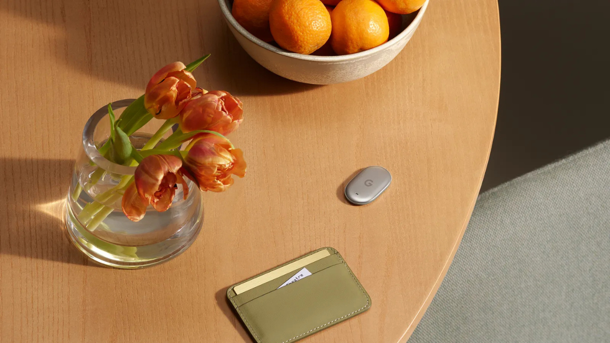 Turn-by-turn directional tracking arrives on the Google Pixel Tag 4 Oval tracker in Fog colour resting on wooden table near green card holder, vase with orange flowers, and fruit bowl lifestyle.