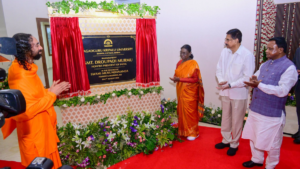 Man in orange robe on left, Jagadguru Kripalu University plaque, woman in orange saree and two men on right clapping cerem...