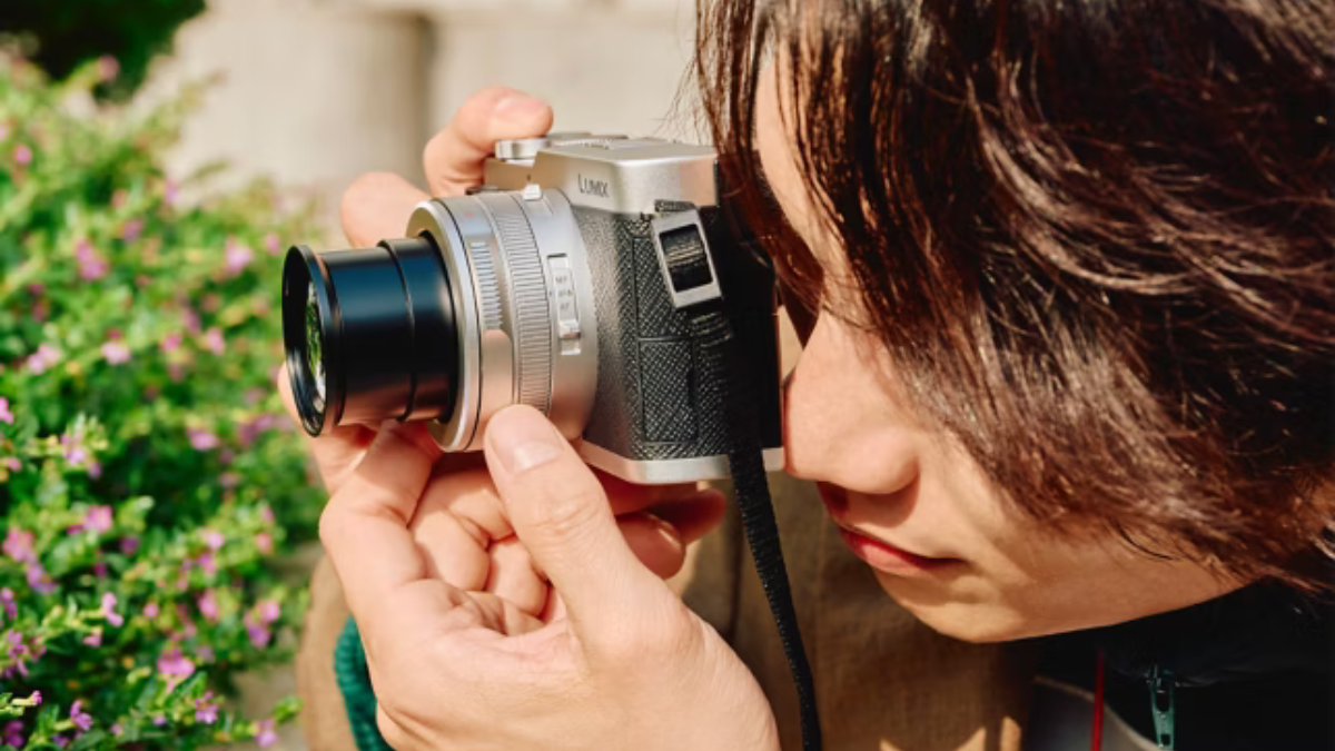 A premium compact camera brings Leica optics to India 1 Person holding a silver compact camera aiming the lens at green foliage and small purple flowers context.