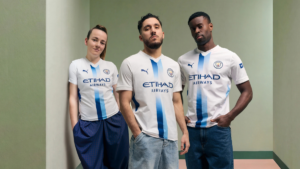 Three models wearing the white and blue Manchester City 26/27 Third Kit standing against a pale green wall indoors.