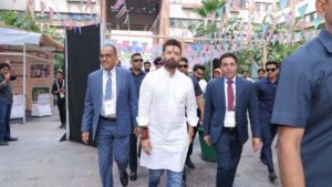 Man in blue suit, man in white kurta, and man in navy suit walking past PMFME Bazaar stalls under colourful flags.