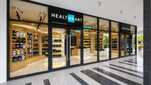 Tiled floor before glass storefront with black frame and HealthKart logo above wooden shelves inside store.