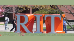 Pedestrians walking past large orange RIT letters on a lawn before a brick building, Bio Frigid Box innovation.