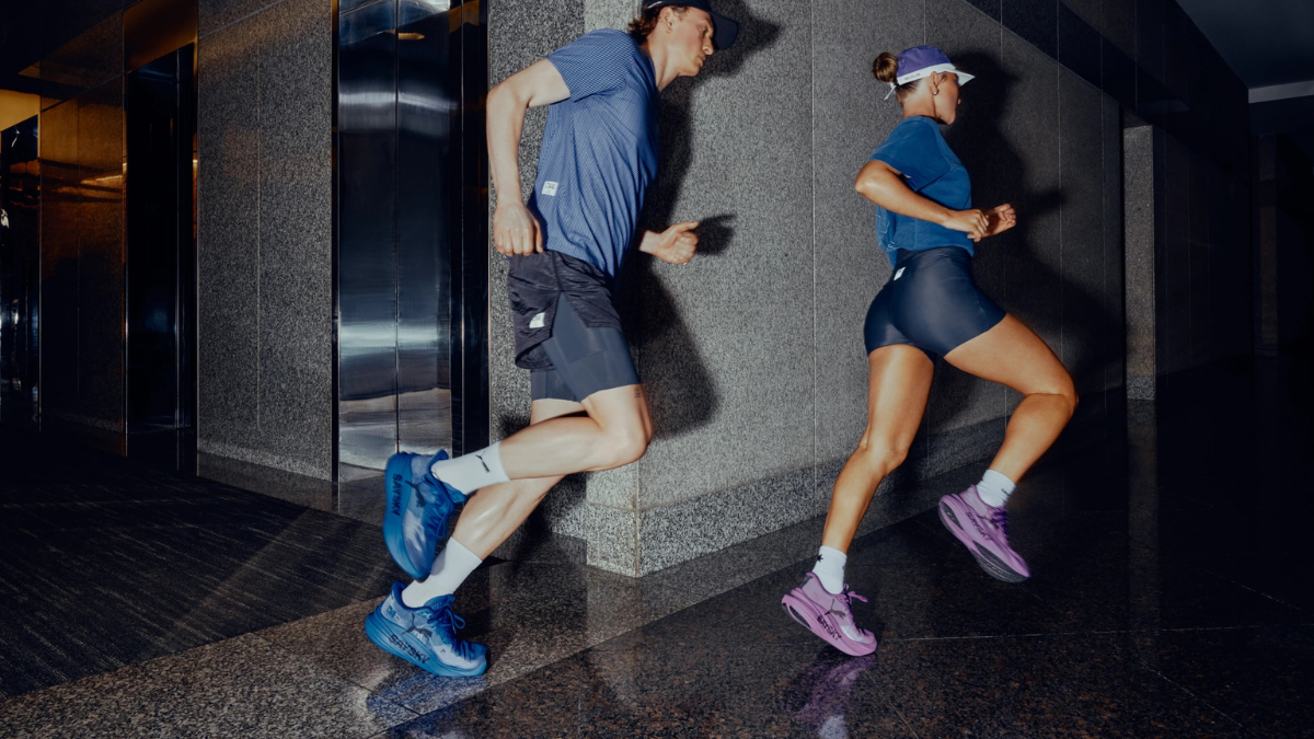 Male runner in blue shoes and female runner in purple shoes sprinting past an elevator along an office corridor.