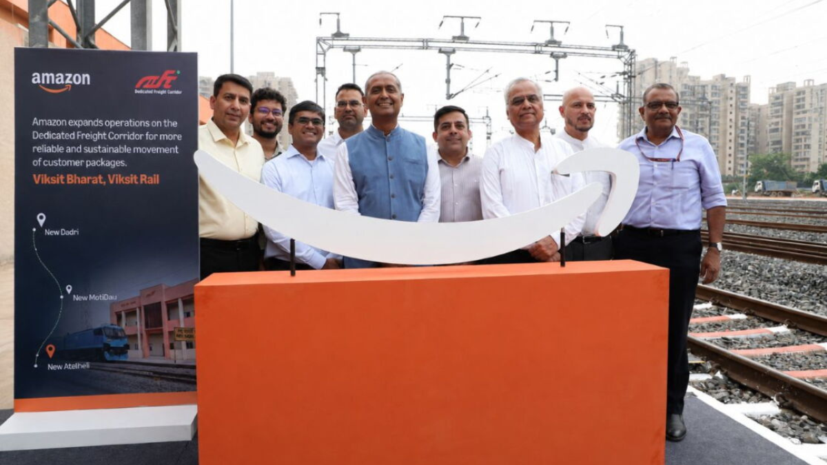 Standing banner on the left beside eight men posed behind a white smile logo on an orange plinth near railway tracks.