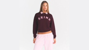 Woman in a maroon SKIMS sweatshirt and light pink sweatpants against a white studio background apparel.