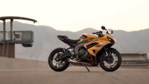 A tower sits on the left with a yellow Triumph Daytona 660 parked on tarmac before hills in the background.