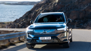 A guardrail and water sit left, with a Volvo EX90 driving forward on a road before a rocky cliff on the right.