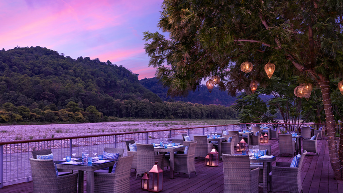 Wicker dining tables with blue cushions, lanterns, hanging lamps, riverside deck railing, riverbed, and forested hills.