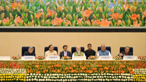 Yellow and orange flowers in foreground, dignitaries seated at a long table during the NGT Mobile Application event.