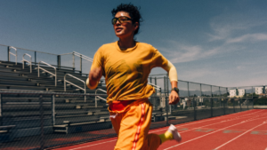 A runner in yellow athletic wear runs forward on a red track, flanked by stadium bleachers on the left and a chain-link fe...