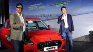 A man stands left of a red Swift giving a thumbs up, with another man on the right before an Auto Green Mission banner.