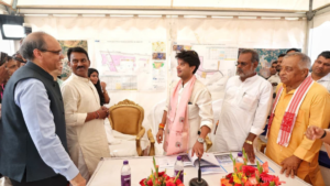 Officials stand beside flowers at a table with Jyotiraditya Scindia in front of Telecom Manufacturing Zone maps.