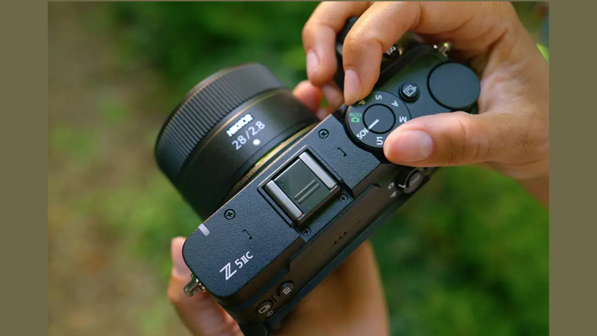 Mounted lens on the left, hands turning the camera top mode dial against blurred foliage background outdoor.