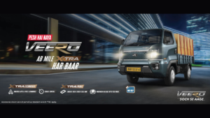 Promotional text sits on the left, with a grey Mahindra Veero truck carrying cargo on the right against city buildings.